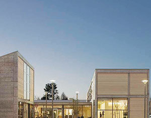 Modern building with large windows and minimalist design, illuminated from within at dusk. The structure has two main sections separated by a walkway, with small shrubs and streetlights in front.