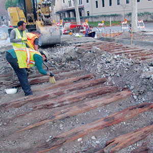 Two construction workers in safety vests examine old wooden beams unearthed at a construction site, with a backhoe and caution barriers in the background.