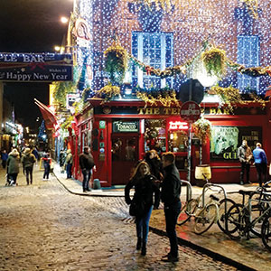 A lively cobblestone street at night with people walking by a brightly lit pub decorated with festive lights and greenery; bicycles are parked outside and the pub’s warm interior glows through the windows.