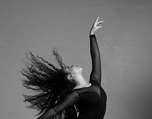 A ballerina in a black tutu and pointe shoes poses gracefully on one foot, with her other leg bent and arms reaching upward. Her long hair flows dramatically as she dances against a plain background.