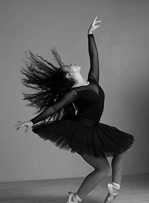 A ballerina in a black tutu and pointe shoes poses gracefully on one foot, with her other leg bent and arms reaching upward. Her long hair flows dramatically as she dances against a plain background.