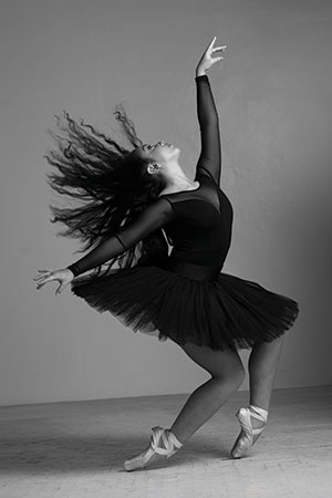 A ballerina in a black tutu and pointe shoes poses gracefully on one foot, with her other leg bent and arms reaching upward. Her long hair flows dramatically as she dances against a plain background.