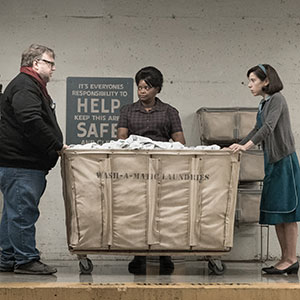 Three people stand around a large laundry cart in front of a wall with a safety sign. Two women, one in uniform, and a man are looking at each other in what seems to be a tense or serious moment.