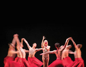 A group of dancers in flowing red skirts move in a circle around a central dancer, who stands still with one arm raised, under dramatic stage lighting against a dark background.