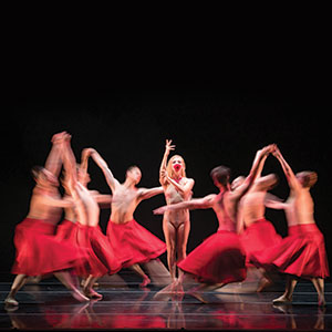 A group of dancers in flowing red skirts move in a circle around a central dancer, who stands still with one arm raised, under dramatic stage lighting against a dark background.