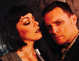A woman with short dark hair and dramatic makeup holds a straight razor close to a serious-looking man’s neck as they stand close together against a dark background.