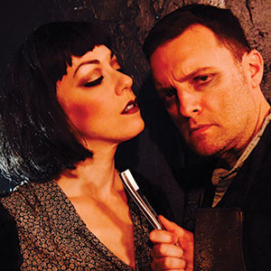 A woman with short dark hair and dramatic makeup holds a straight razor close to a serious-looking man’s neck as they stand close together against a dark background.