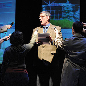 A man in a suit speaks while holding papers on stage as several people surround him, each holding a microphone toward him. Background images are projected onto screens behind them.