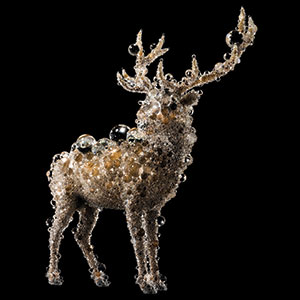 A sculpture of a stag covered in sparkling beads, crystals, and bubbles, standing against a black background. The intricate details give the appearance of the animal being made entirely out of shimmering materials.