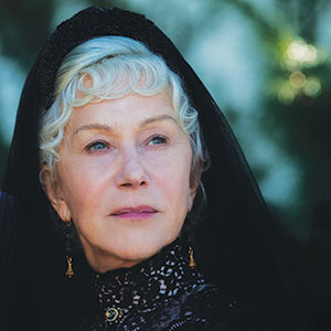 An older woman with short, curly white hair wears a black lace dress, gold earrings, and a black veil, gazing thoughtfully into the distance against a blurred outdoor background.