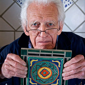 An elderly man with white hair and glasses holds up a colorful, geometric patterned tile. The background features light-colored tiles arranged in a diamond pattern.