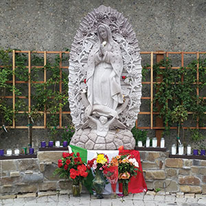 A stone statue of the Virgin Mary stands on a pedestal, surrounded by flowers, candles, and an arrangement of the Mexican flag at the base. The background features a trellis with greenery on a stone wall.
