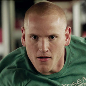 A man with a shaved head and intense expression looks forward while wearing a green shirt. The background is blurred, suggesting an indoor setting, possibly a gym or sports facility.