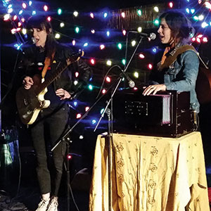 Two women perform on stage, one playing guitar and singing, the other singing and playing a keyboard instrument on a table, with colorful string lights in the background.