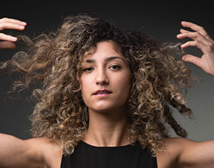 A person with curly, light brown hair wearing a sleeveless black top lifts their arms to touch their hair, looking confidently at the camera against a dark, neutral background.