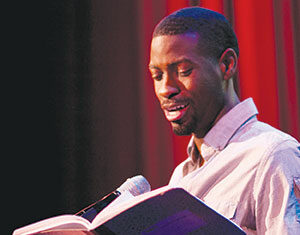A man in a light shirt stands at a microphone, reading aloud from an open book, with red curtains in the background.