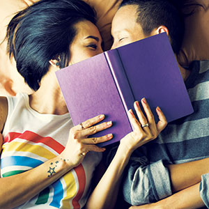 Two people lie side by side on a bed, holding a purple book that partially hides their faces. One person has a rainbow shirt and visible tattoos. They appear to be smiling and sharing a tender, intimate moment.