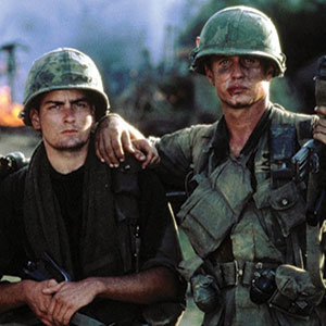 Two soldiers in military uniforms and helmets stand side by side, one with his arm around the others shoulder. They have serious expressions and appear to be in a combat zone, with smoke and fire in the background.