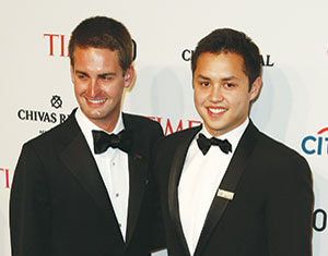 Two men in black tuxedos and bow ties smile while posing together on a red carpet at a formal event, with branded sponsor logos and a TIME backdrop behind them.