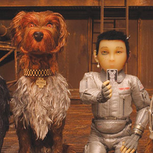 A dog with shaggy brown fur and a collar labeled KING sits next to a person in a silver spacesuit, who is holding up a small black-and-white object. Both are facing forward.