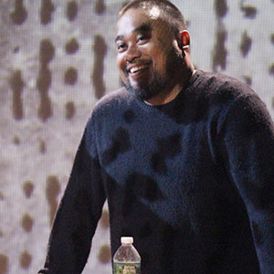 A man with short hair and a beard, wearing a black sweater, smiles while standing behind a table with a water bottle on it. The background is blurred with abstract patterns.