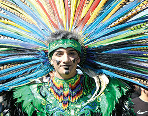 A person wearing a vibrant, feathered headdress and green, colorful traditional attire stands outdoors, smiling. The headdress features long, multicolored feathers radiating outward.