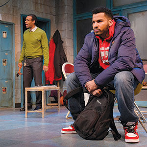 A young man in a red hoodie and blue jacket sits on a chair holding a black backpack, looking serious. Behind him, another man in a green sweater stands near blue lockers, appearing to speak or shout.