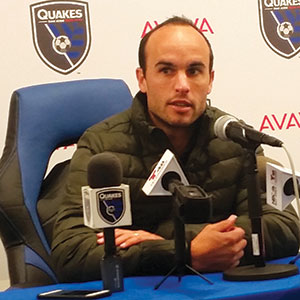 A man in a green jacket sits at a press conference table, speaking into microphones branded with the San Jose Earthquakes soccer teams logo, with a team logo and AVAYA in the background.