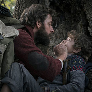 A bearded man covers a young boys mouth with his hand as they hide near a tree, both looking tense and scared. The man wears a red sweater and backpack; the boy wears a patterned sweater.
