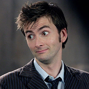 A man with tousled brown hair wearing a pinstripe suit and tie looks to the side with a raised eyebrow and a slightly amused or skeptical expression.