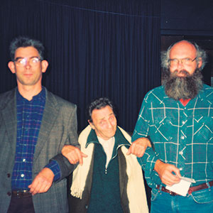 Three men stand together indoors against a dark curtain. The man in the middle wears a light-colored scarf and jacket, flanked by two men; one in glasses and a blazer, the other with a long beard and checkered shirt.
