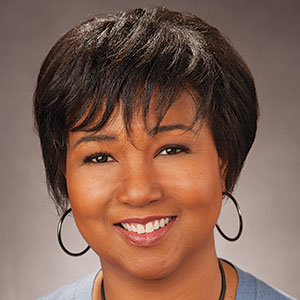 Smiling woman with short dark hair, hoop earrings, and a light blue top, photographed against a neutral brown background.