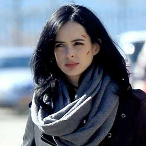 Woman with long black hair wearing a large gray scarf and dark jacket, standing outdoors with a slightly serious expression and a few scratches on her face.