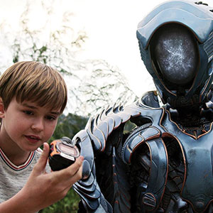 A young boy in a striped shirt shows a device to a tall, futuristic robot with a smooth, featureless face and metallic armor, outdoors with greenery in the background.