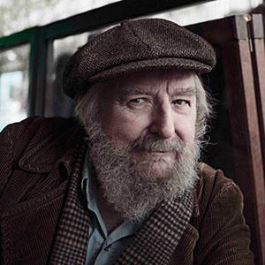 An older man with a thick gray beard and mustache, wearing a brown flat cap and brown corduroy jacket, sits indoors and smiles gently at the camera.