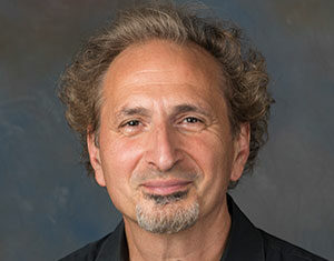 A middle-aged man with curly, light brown hair, a goatee, and a warm smile wearing a black collared shirt, posed against a neutral, softly blurred background.