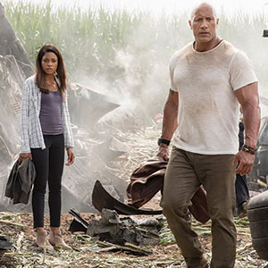 A man in a dirty white t-shirt and a woman in casual clothes stand among debris in a field, with smoke rising in the background and tall corn plants behind them. Both appear alert and tense.
