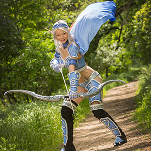 A person in a fantasy archer costume with elf ears, silver wig, and blue armor poses outdoors on a sunlit forest path, holding a bow and smiling.