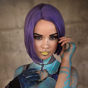 A person with a purple bob wig and creative face and body paint resembling a futuristic or sci-fi character poses with a hand near their face. They wear makeup with yellow lips and blue, black, and yellow designs.