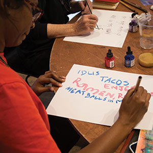 Two people sit at a round table, painting words on paper. One person writes colorful words like TACOS, Ramen, and MEATBALLS IN N.Y. with paintbrushes; paint bottles and supplies are on the table.