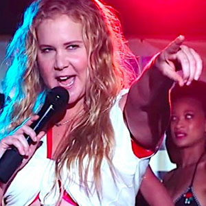 A woman with long blonde hair holds a microphone and points forward while singing on stage, with colorful lighting and another person visible in the background.