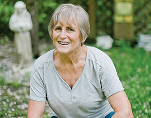 Older woman with short gray hair, wearing a light gray shirt and blue jeans, squatting and smiling outdoors in a grassy garden with a statue in the background.