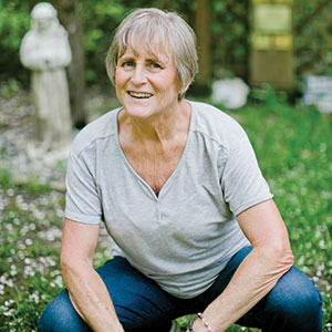 Older woman with short gray hair, wearing a light gray shirt and blue jeans, squatting and smiling outdoors in a grassy garden with a statue in the background.