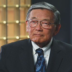 An older man with short gray hair and glasses, wearing a dark pinstripe suit, white shirt, and blue patterned tie, sits in front of a softly lit background with square gold accents.