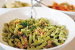 A close-up of a bowl of green pasta garnished with chopped herbs and grated cheese, with other dishes slightly blurred in the background.