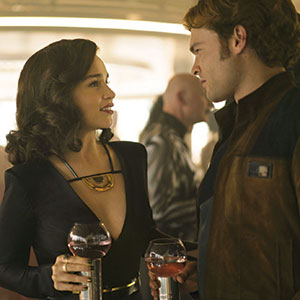 A woman and a man stand close together in a futuristic setting, each holding a glass of red drink. The woman wears a dark dress and gold necklace, while the man wears a brown jacket. They appear to be having a conversation.