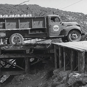 A vintage dump truck with SOC on the door drives over a wooden bridge in a mountainous area. The bridge appears unfinished or in disrepair, and the truck is partially on the edge.
