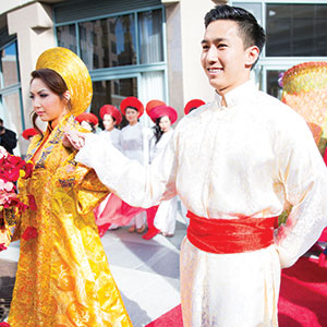 A bride and groom in traditional Vietnamese wedding attire, with the bride in a gold áo dài and headdress, and the groom in a white outfit with a red sash, walking outside with attendants in the background.