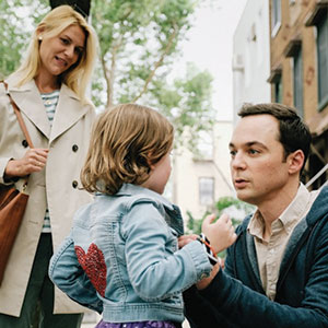 A man crouches to talk to a young girl wearing a denim jacket with a red heart on the back, while a woman stands nearby, smiling. They are outdoors on a city street with trees and buildings in the background.