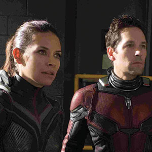 Two people wearing red and black superhero suits stand side by side, looking intently ahead. The background is dimly lit, with part of a wall and some yellow railing visible.